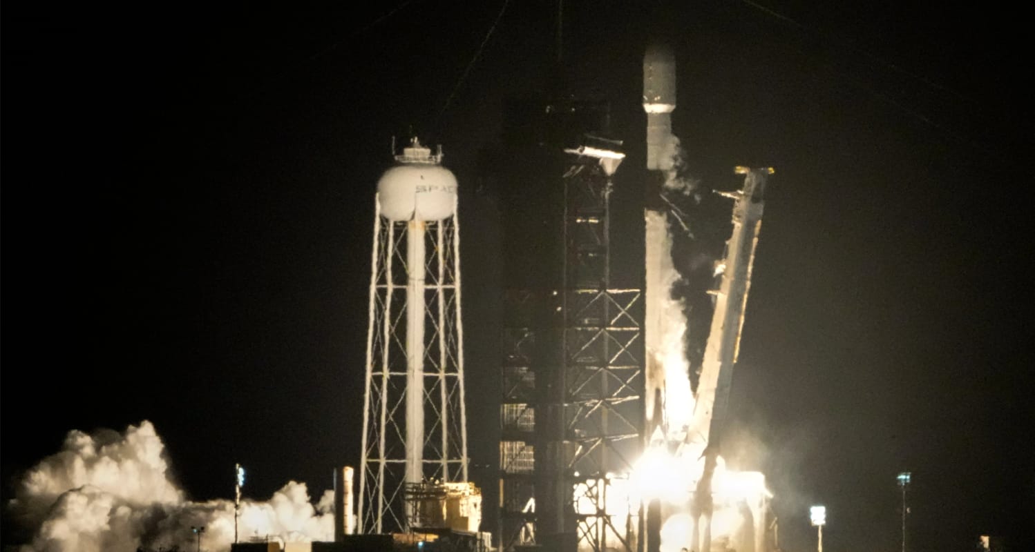 A rocket blasts off from the ground at night aside a water-tower looking structure. 